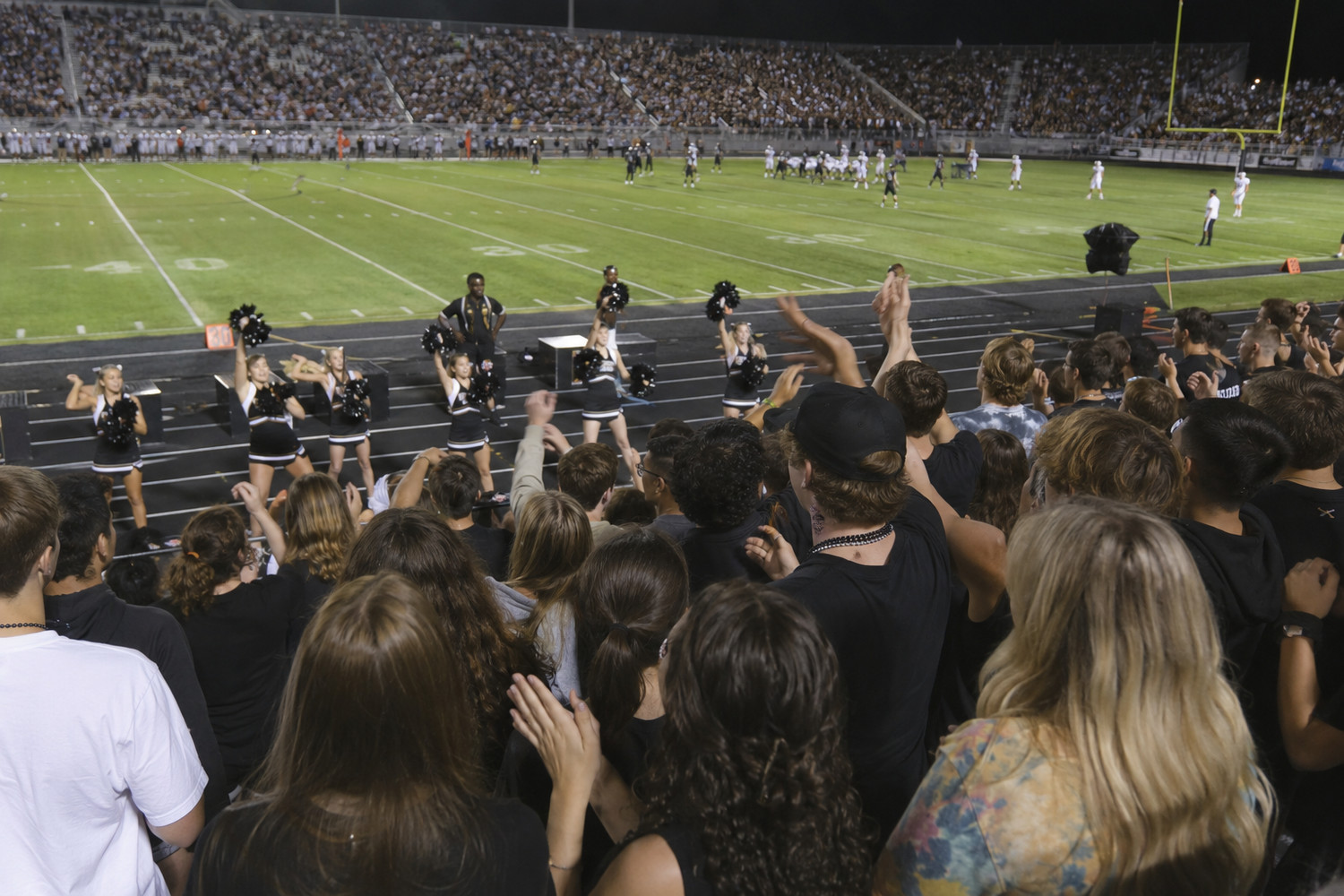 Crowd of fans in black attire cheering at a nighttime high school football game in a packed stadium.