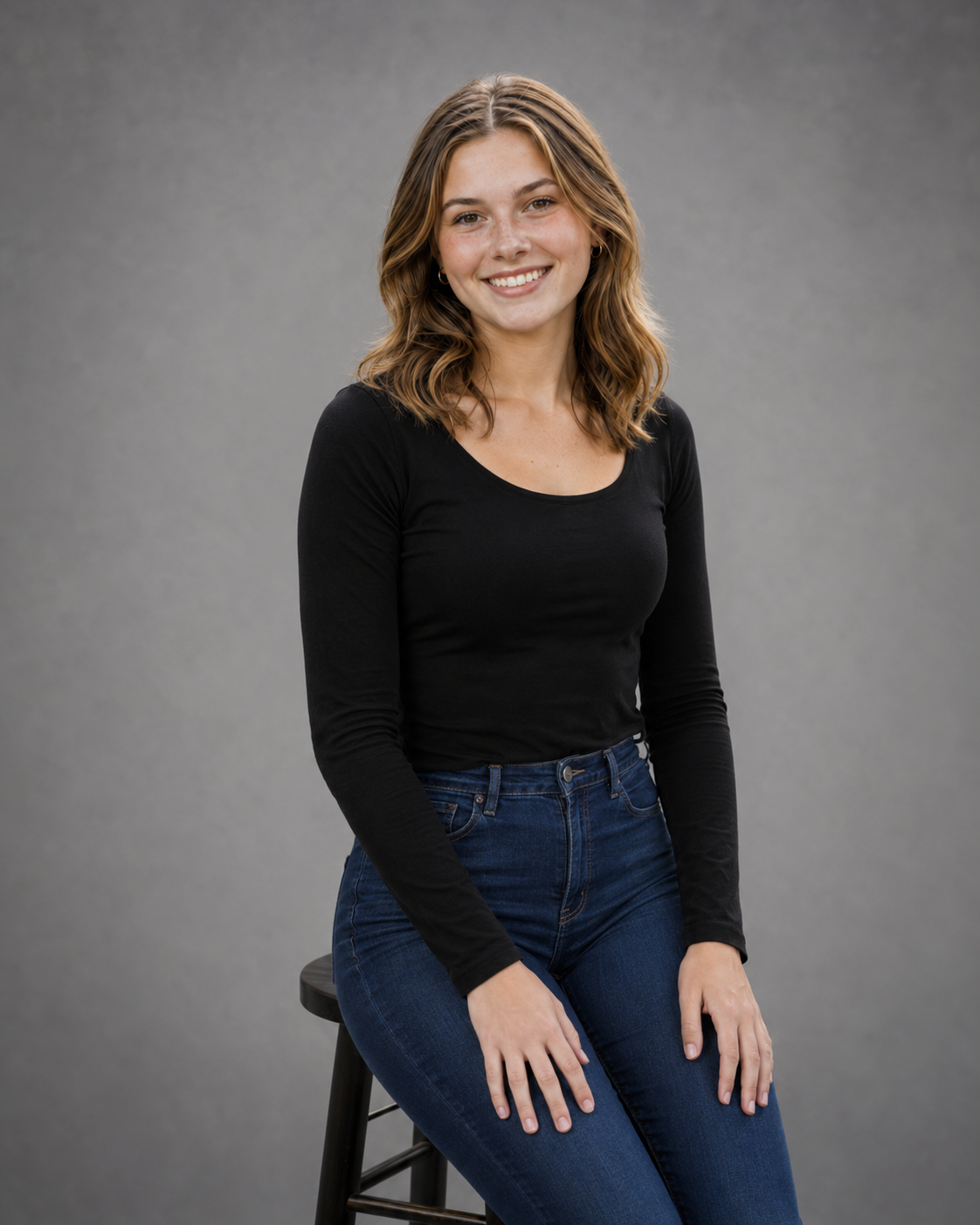 Smiling young woman in black long-sleeve top and jeans sitting on stool against gray studio background.