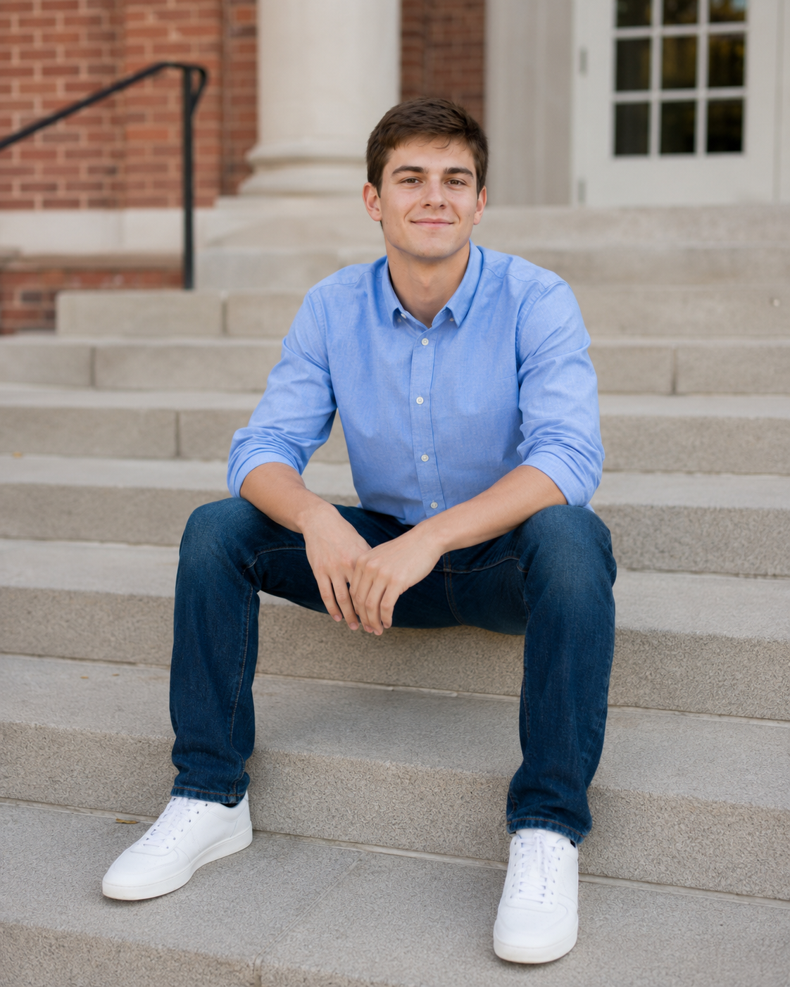 Young man in blue shirt and jeans sitting on university steps smiling confidently outdoors.