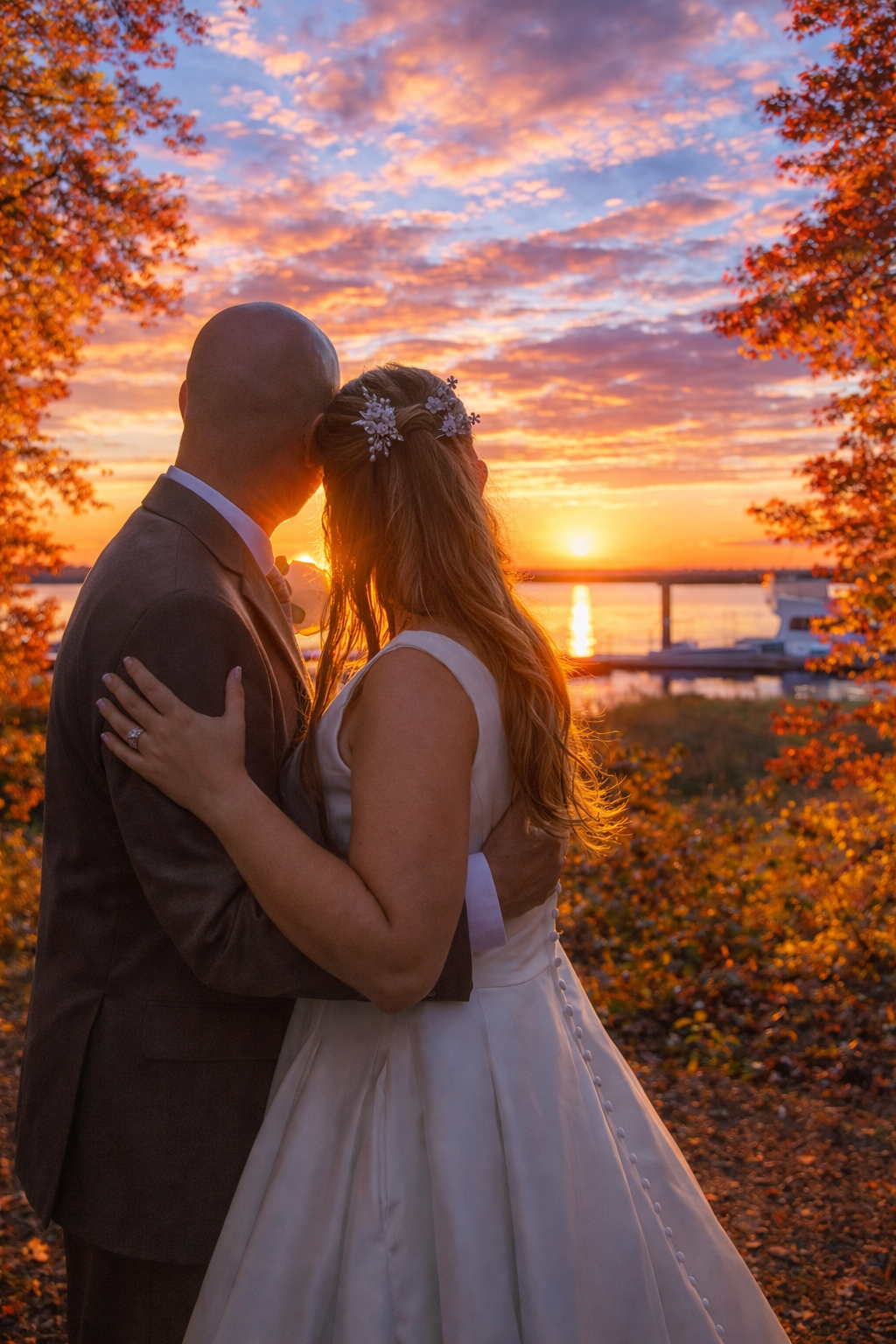 Bride and groom embrace at sunset by a waterfront, framed by autumn foliage under a vibrant orange sky.