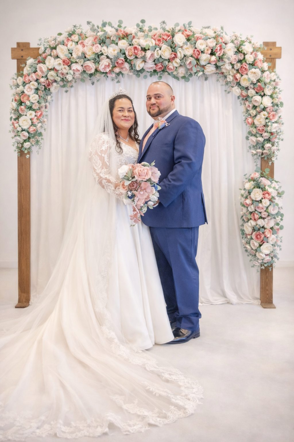Bride and groom posing together before pink floral arch, she in white gown, he in blue suit.