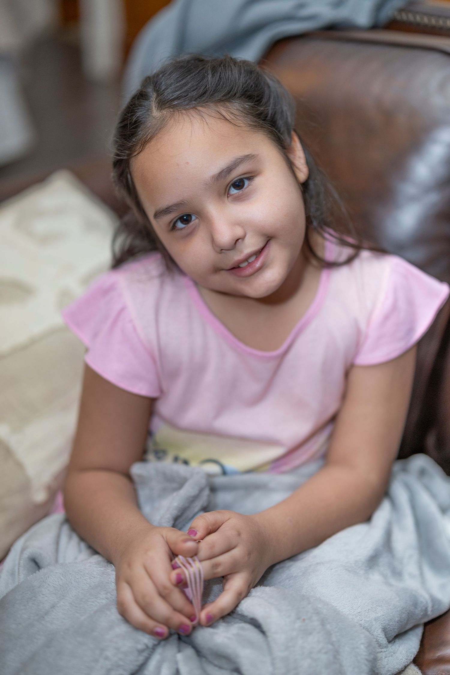 Child in a pink t-shirt sitting comfortably on a leather chair with a grey blanket.