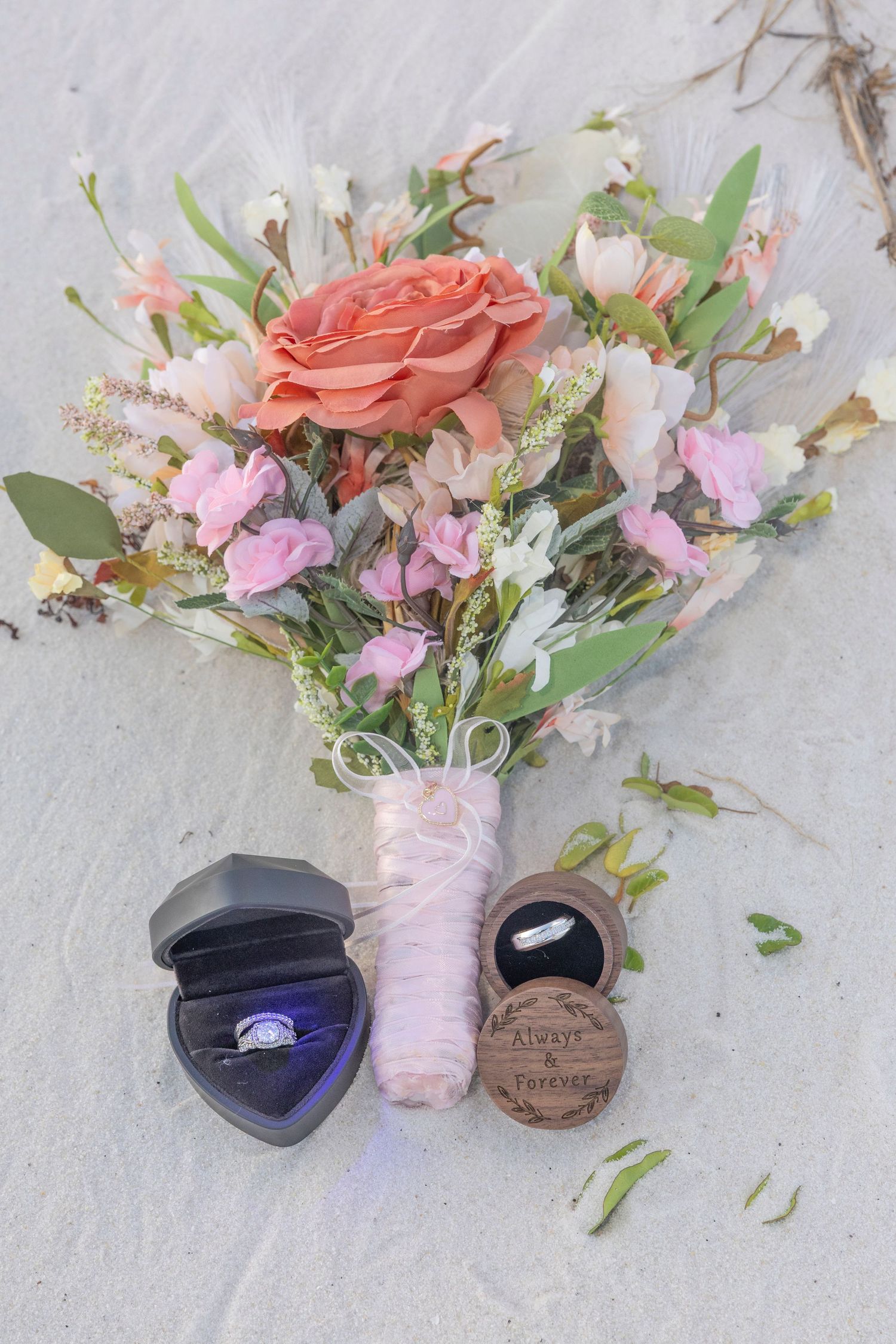 Wedding bouquet with coral rose and pink flowers alongside ring box and wooden ring holder on white textured surface.