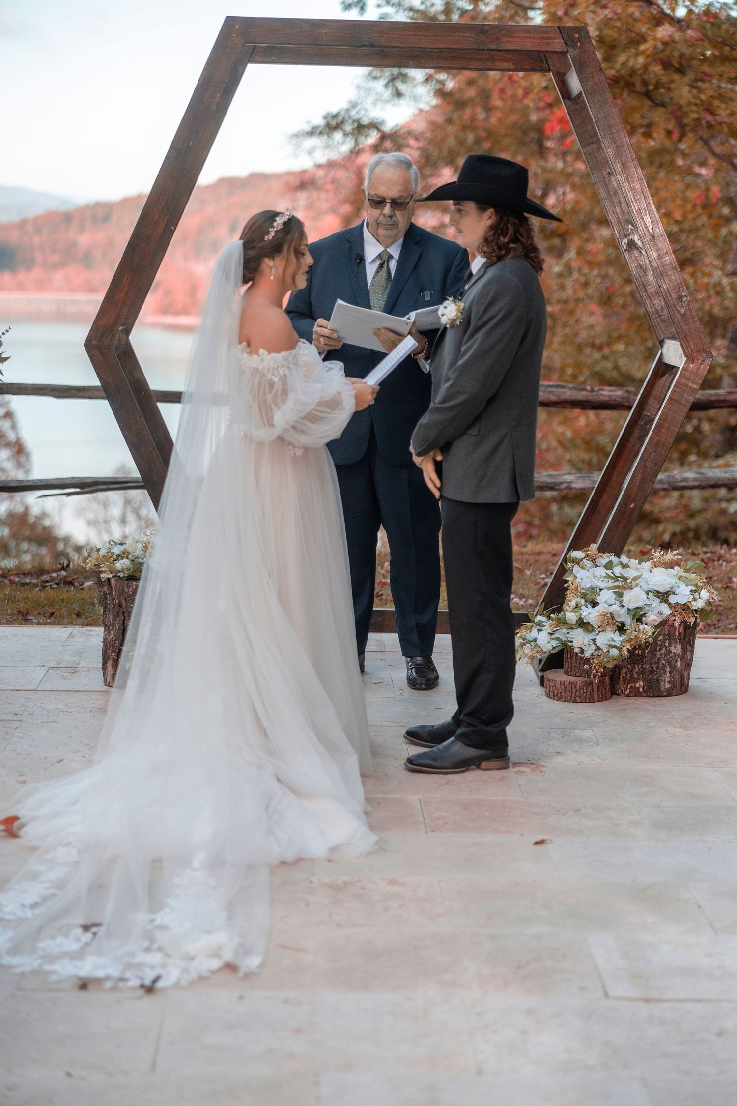 Outdoor wedding ceremony under autumn trees with bride in flowing white gown and officiant in dark attire.
