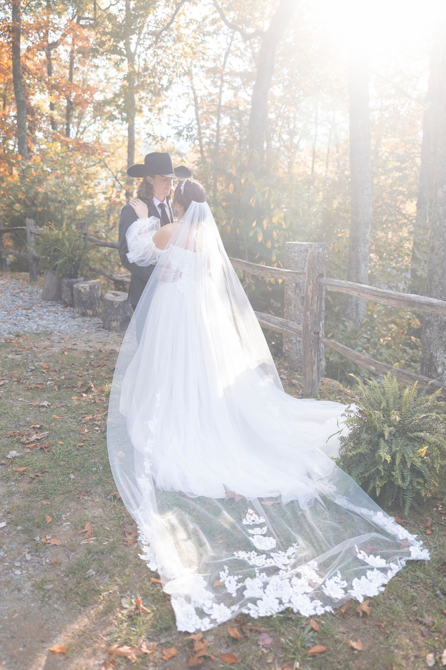 A couple embraces in an autumn forest setting with a flowing white wedding dress and veil trailing on fallen leaves.