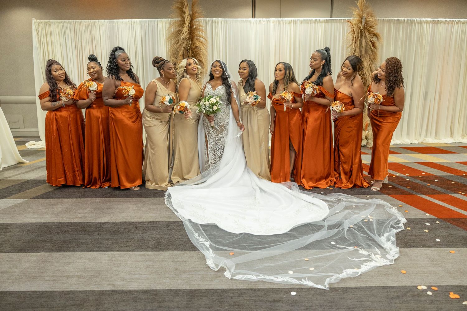 A wedding party photo showing bridesmaids in orange dresses lined up with the bride in a flowing white gown with long train.