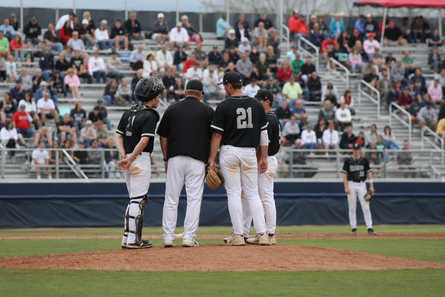 Baseball coach visits pitcher on mound during game with catcher and teammates gathered.