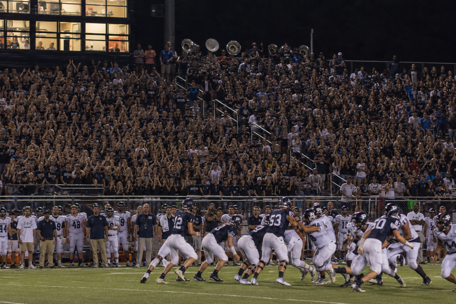 College football game with packed stadium crowd cheering as players compete on the field under bright lights at night.