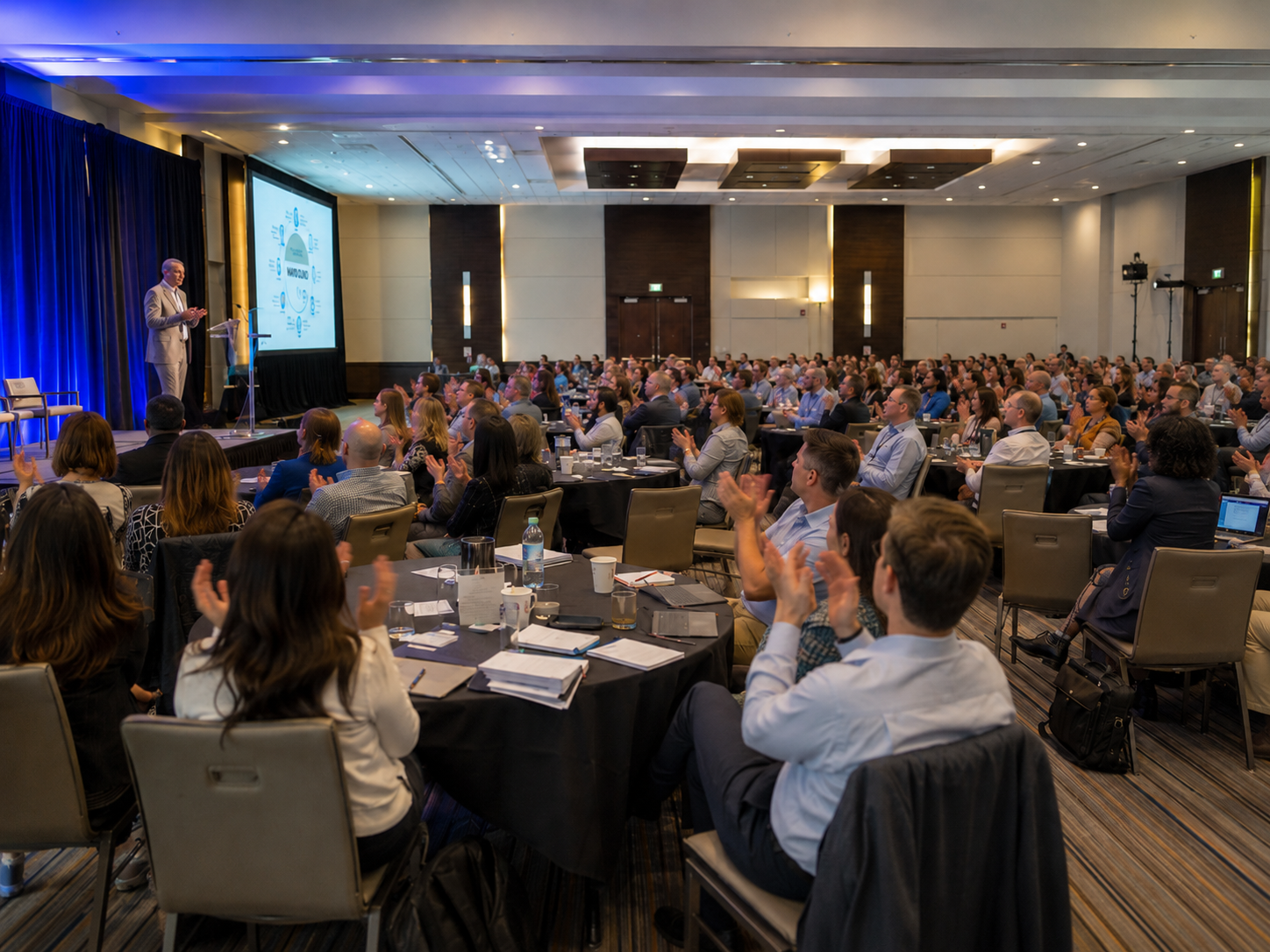 Large audience applauding a speaker presenting on stage at a professional business conference event.