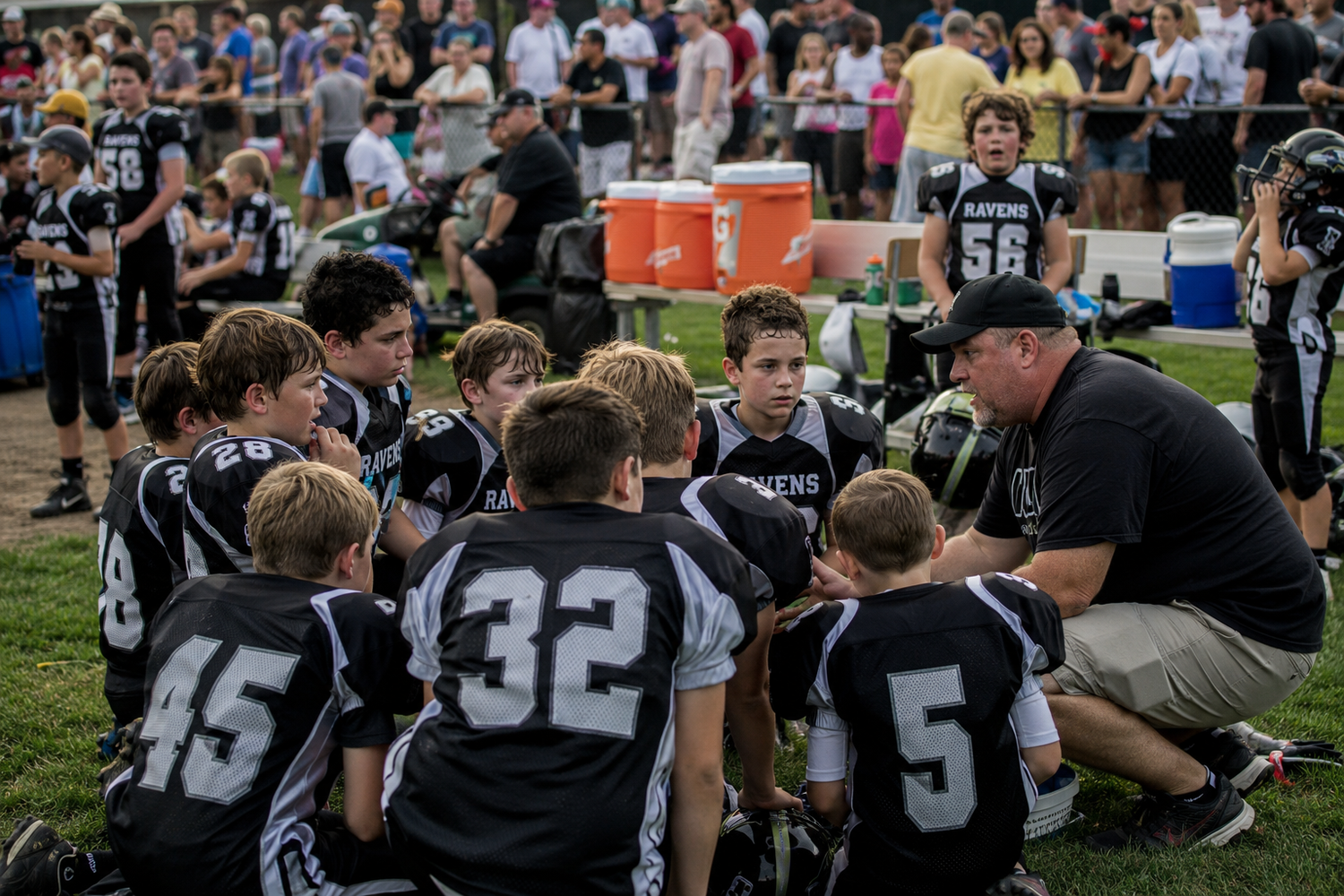 Youth football team huddles with coach on sideline during a game, players wearing black uniforms.