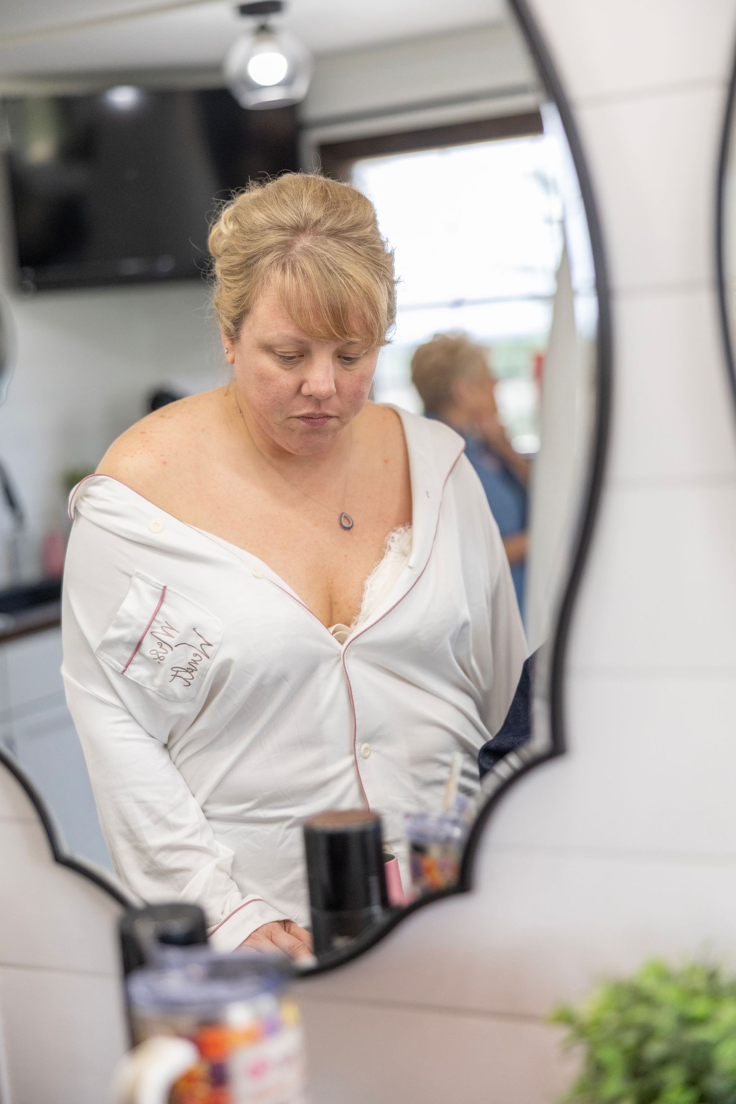 A blonde woman in a white robe looks down while standing at a bathroom counter, reflected in an ornate mirror.
