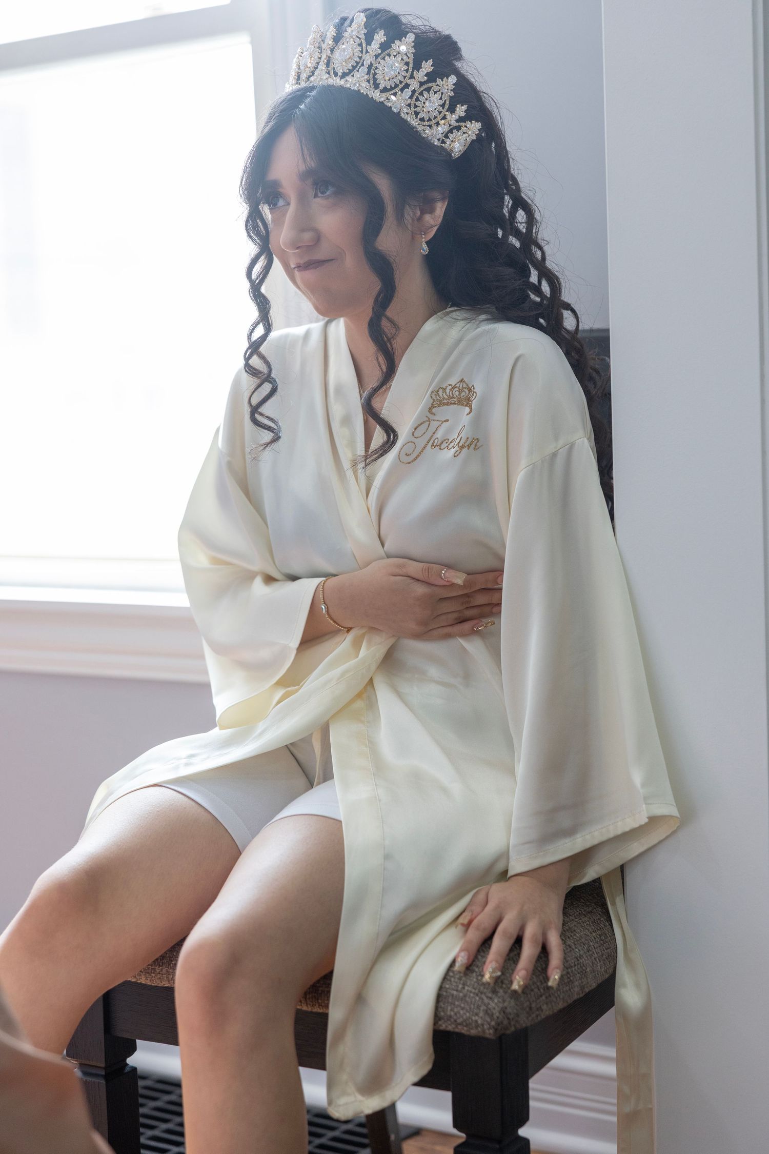 Bride wearing a white robe and crystal tiara sits gracefully on a chair near a bright window.