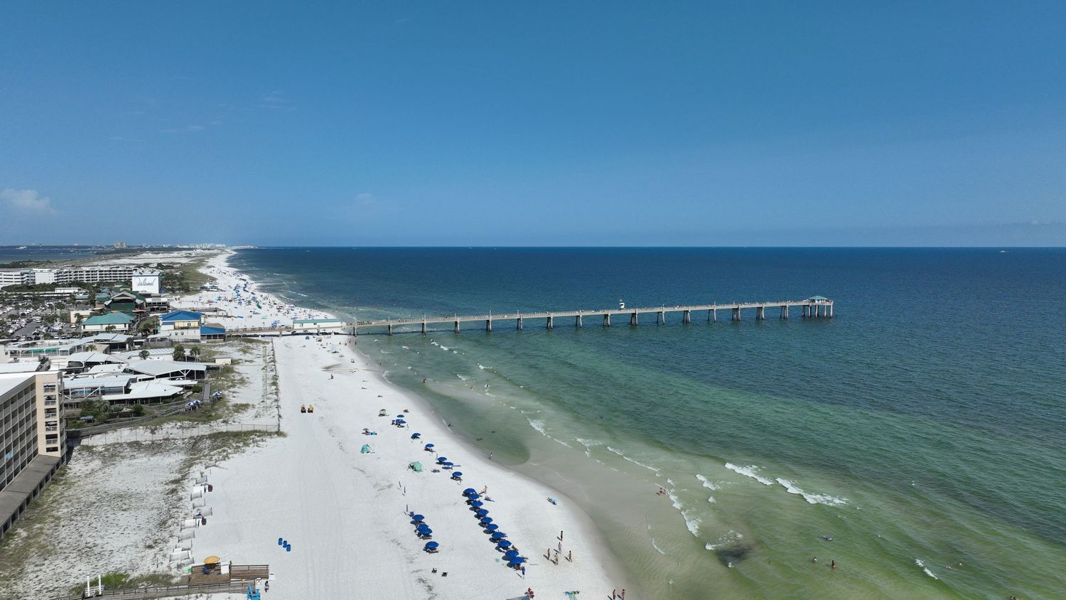 Aerial view of a white sandy beach with a fishing pier extending into clear turquoise Gulf waters under a bright blue sky.