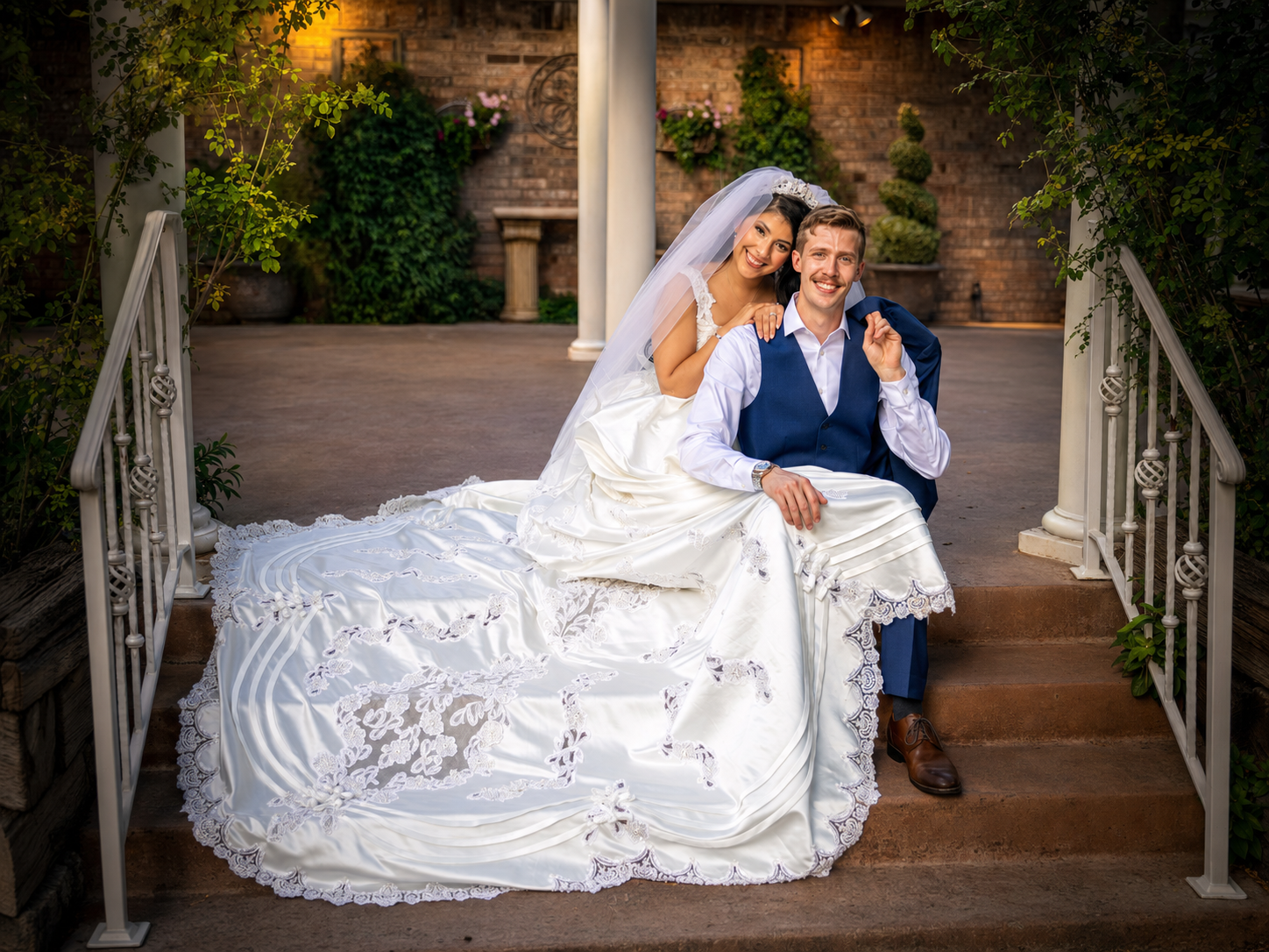 Bride in elegant white gown and groom in blue suit pose on stone steps at an outdoor wedding venue.