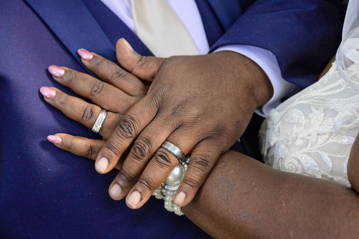 Newlywed couple displaying wedding rings on their hands, bride with pink nails wearing pearl ring beside groom in blue suit.