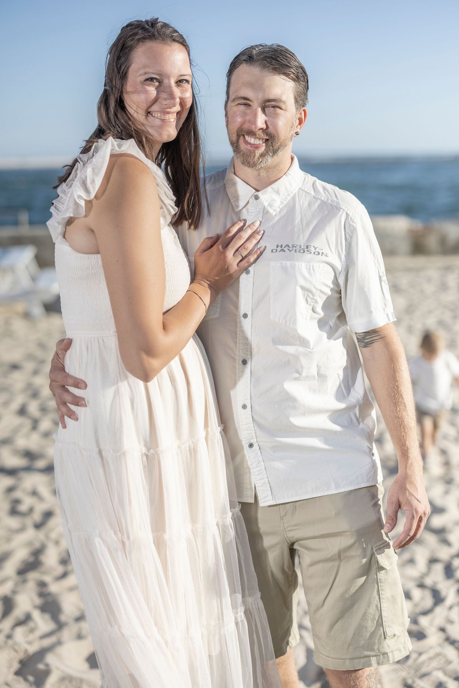 A couple in light summer attire poses together on a sunny beach.
