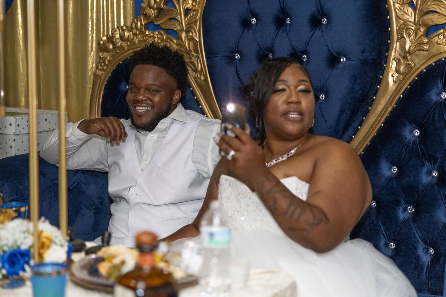 A couple in wedding attire sits on ornate blue and gold thrones at their wedding reception celebration.