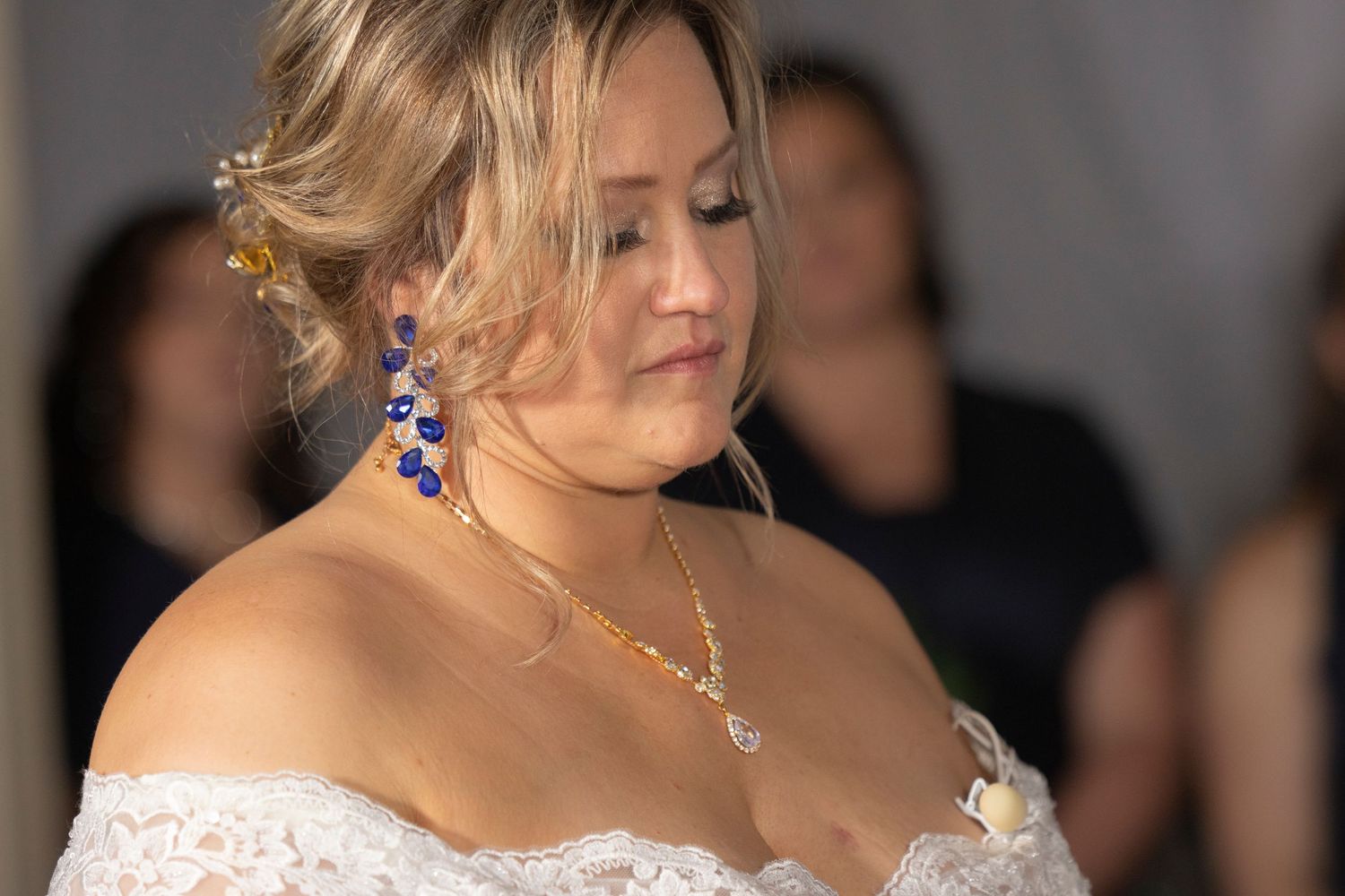 Elegant bride in white lace dress with blue beaded earrings and gold necklace at wedding ceremony.