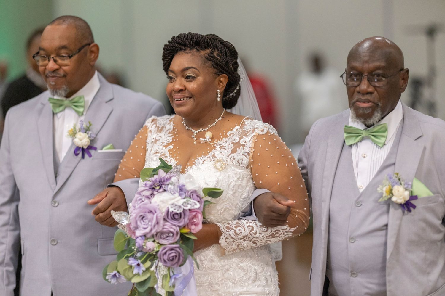 Bride in lace wedding gown holds purple bouquet, flanked by two men in gray suits with green bow ties at wedding ceremony.