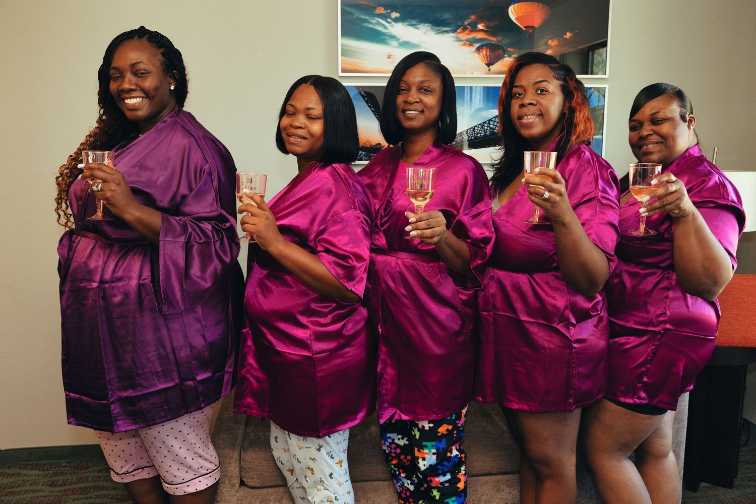 Five women in matching magenta satin robes smile and raise champagne glasses together in a celebratory bridal party photo.