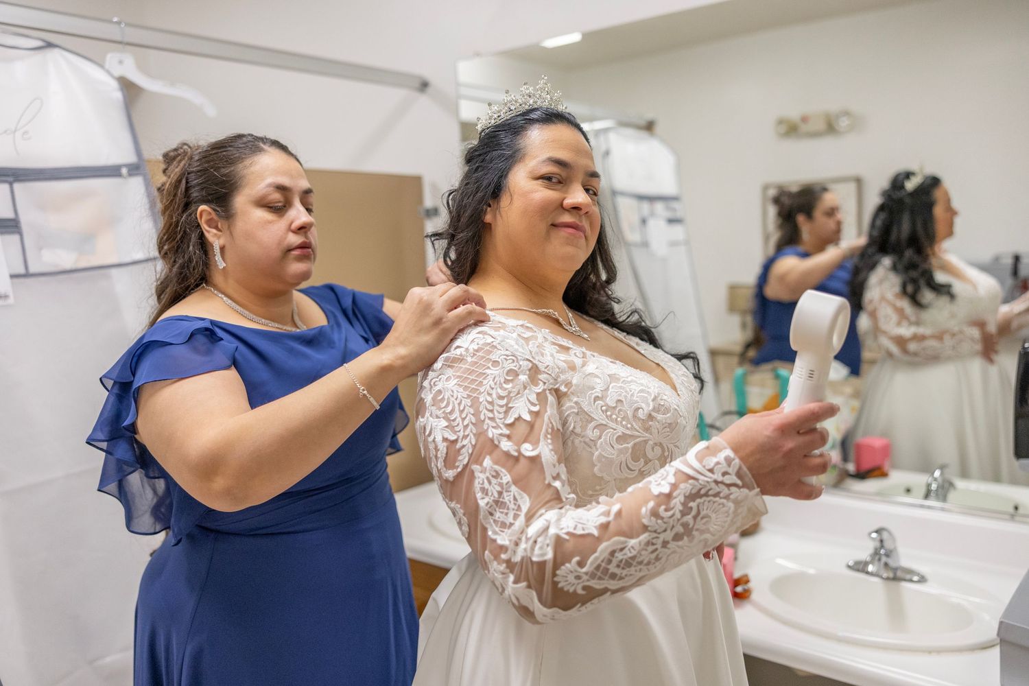 A bridesmaid in blue helps a bride in lace wedding gown prepare in a bathroom mirror setting.