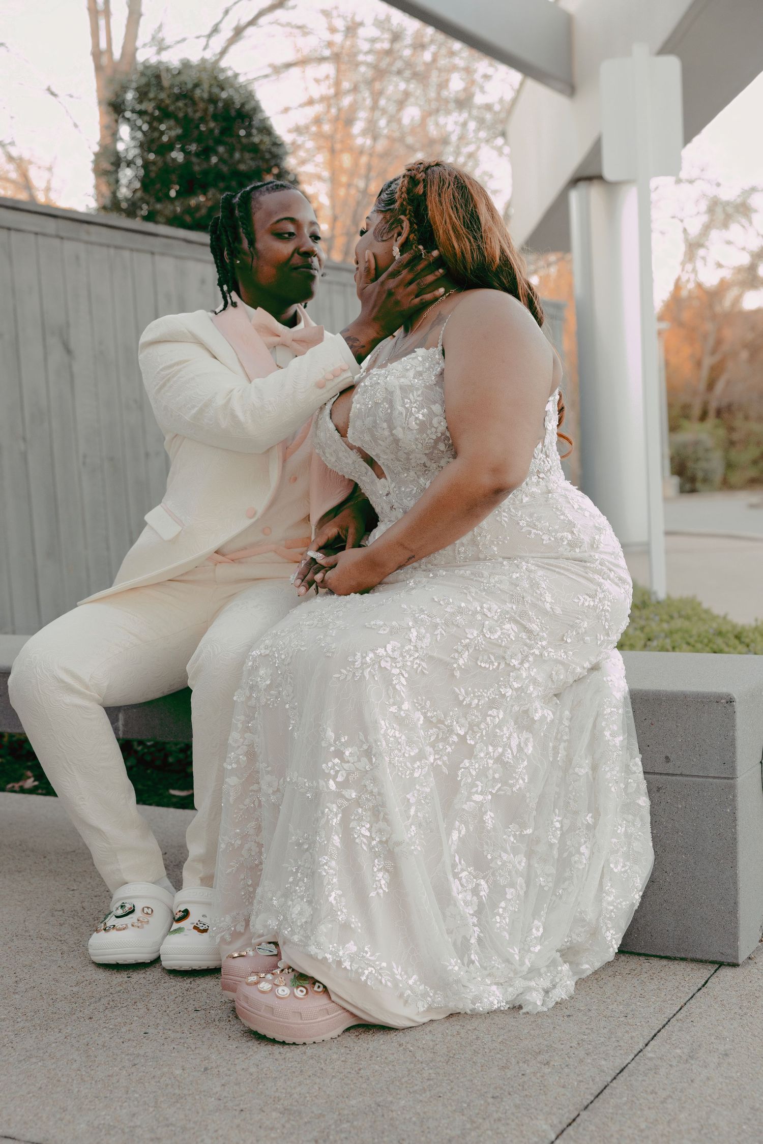 A couple in wedding attire sits together outdoors, the groom in cream suit tenderly holds the bride's face in a romantic moment.