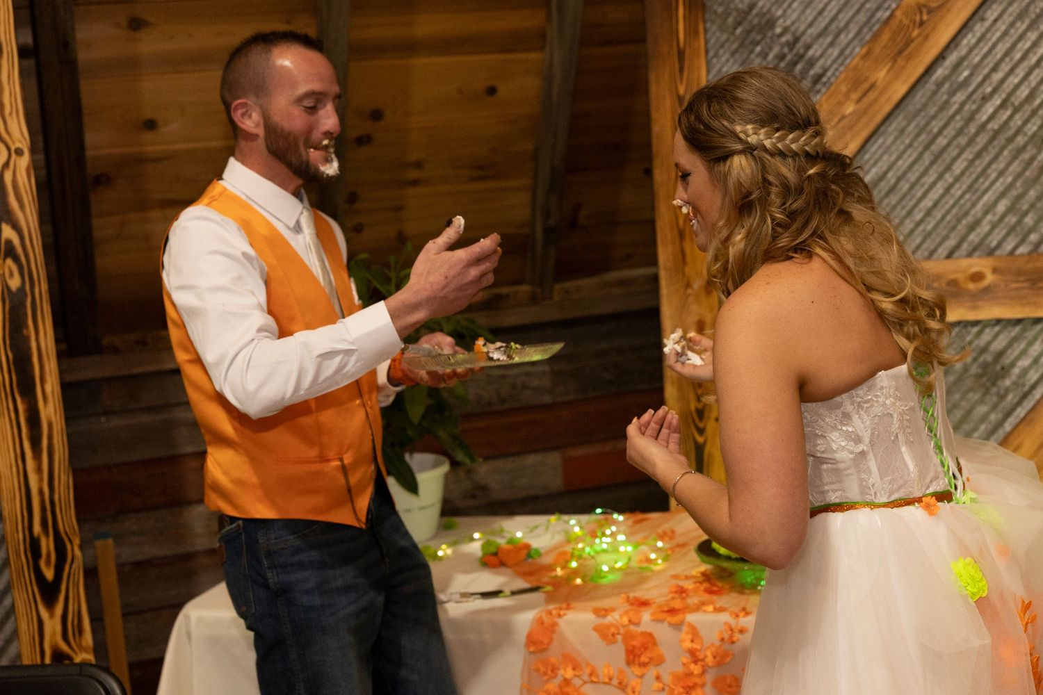 Groom in orange vest and bride in white dress share a joyful moment at their rustic barn wedding reception.