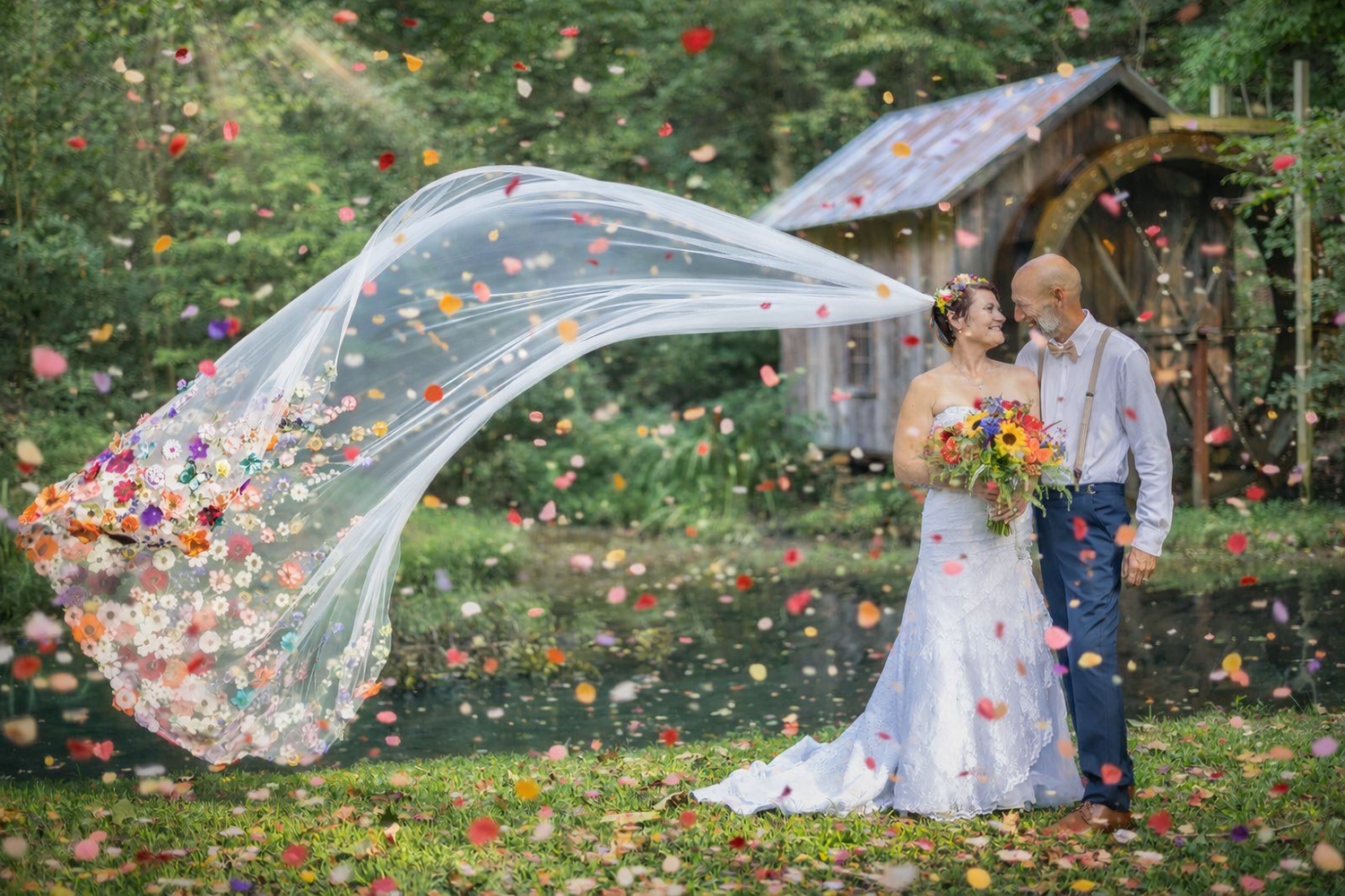 Bride and groom stand outdoors as colorful flower petals and a flowing veil fill the air near a rustic wooden barn.