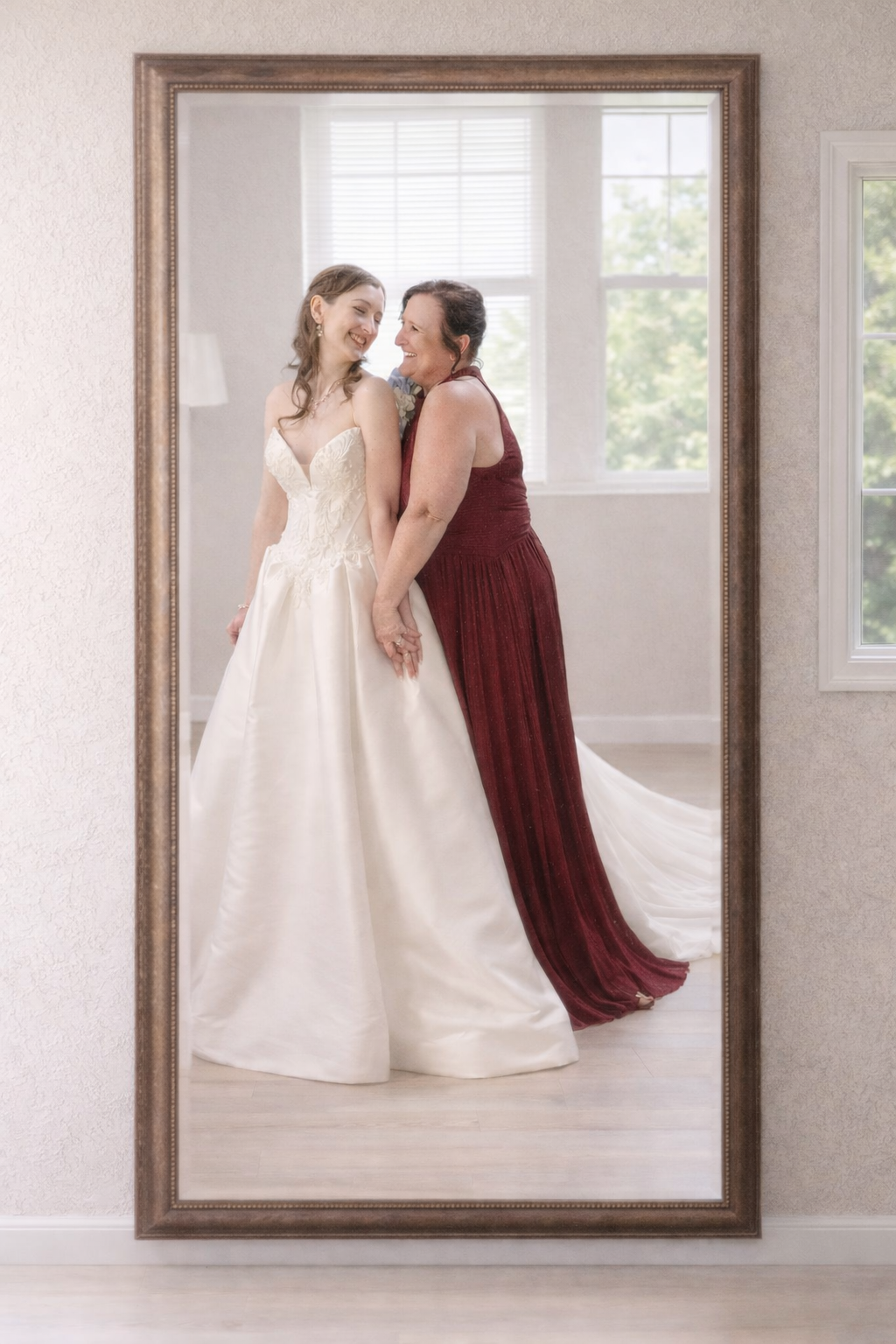 Bride in white ball gown smiles with woman in burgundy dress captured in large ornate mirror.