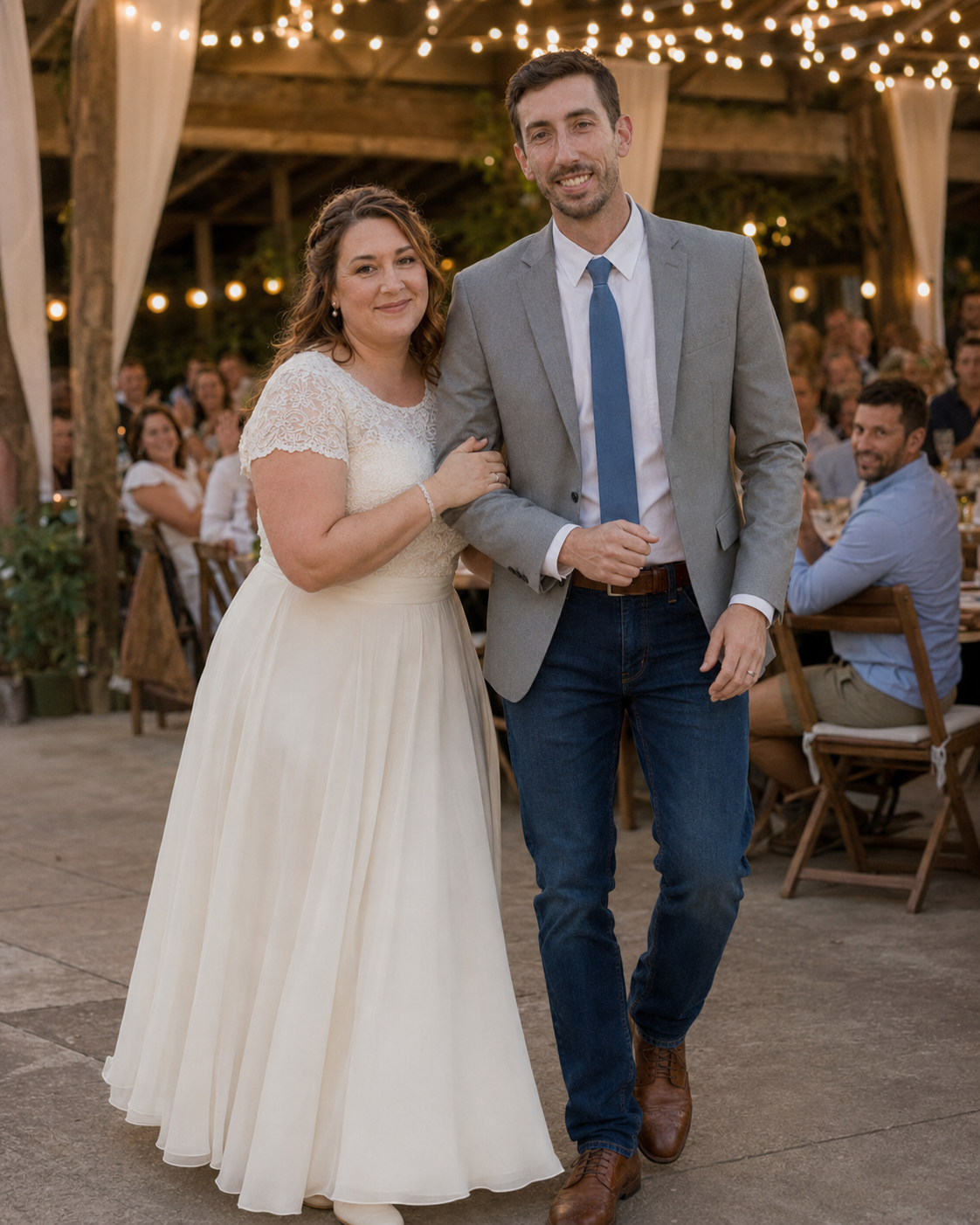 Bride in white lace dress and groom in gray blazer with blue tie pose together at elegant barn wedding reception.