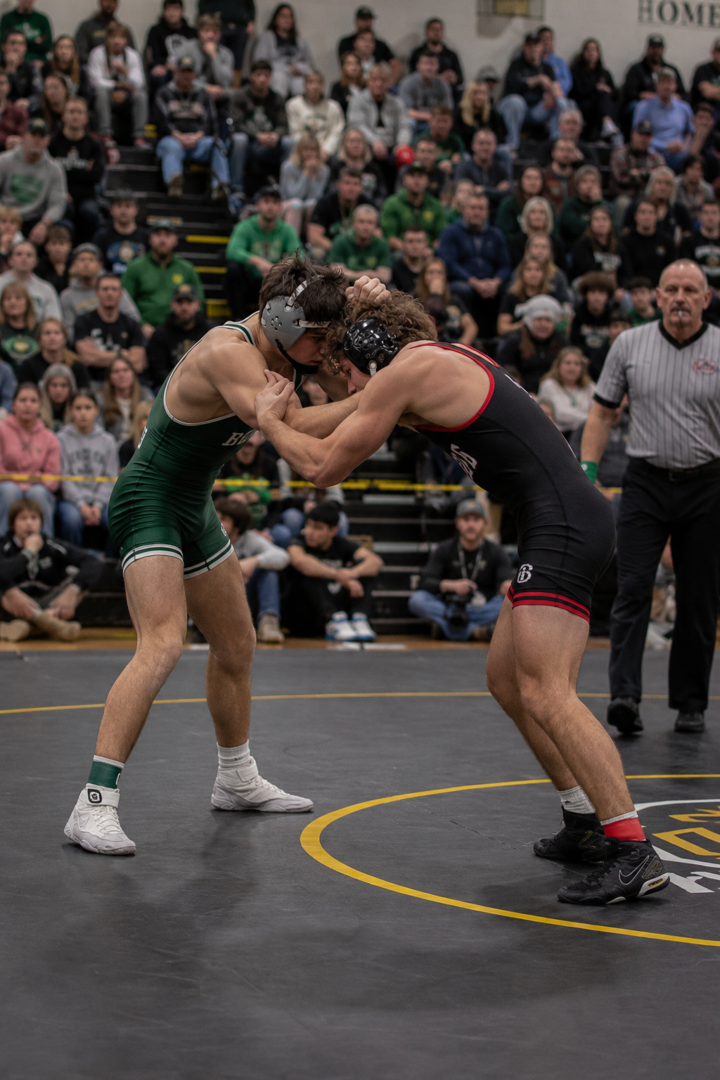 Two wrestlers compete on mat in packed gymnasium during high school wrestling match.