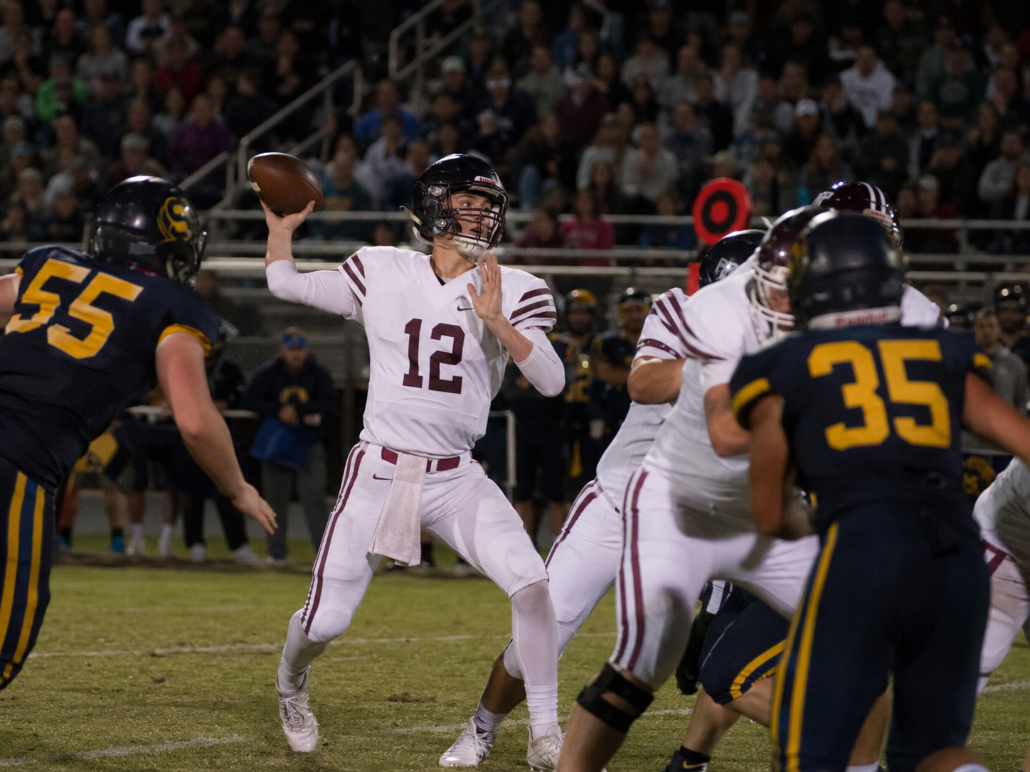 High school quarterback number 12 in maroon throws a pass under pressure during a Friday night football game.