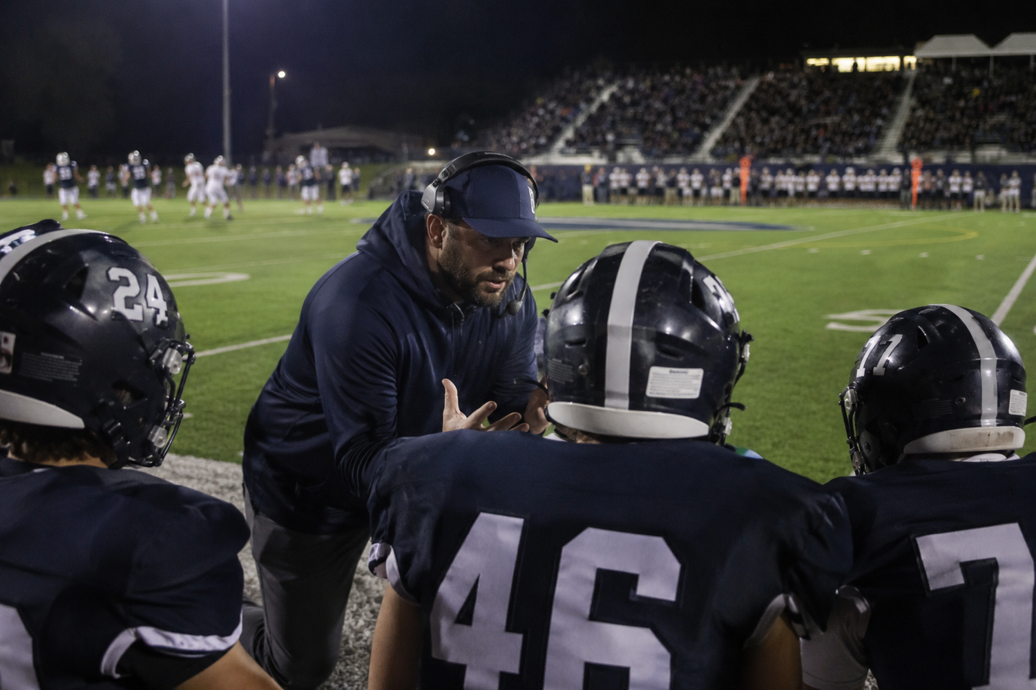 Football coach in navy hoodie reviews plays with players wearing #46 and #7 jerseys during a night game.