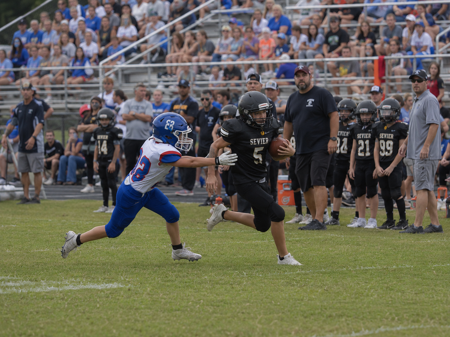 Youth football player number 5 in black uniform stiff-arms a defender during a sideline run.