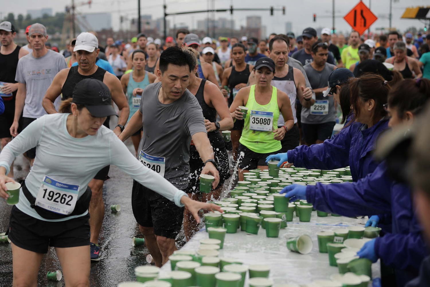 Marathon runners grabbing water cups at a hydration station during a road race, volunteers handing out drinks.