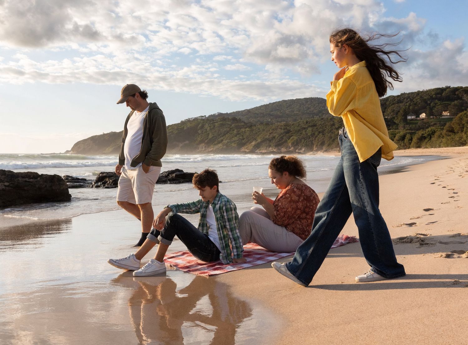 Four friends enjoy a sunny beach day, two sitting on a blanket while others walk along the shoreline.