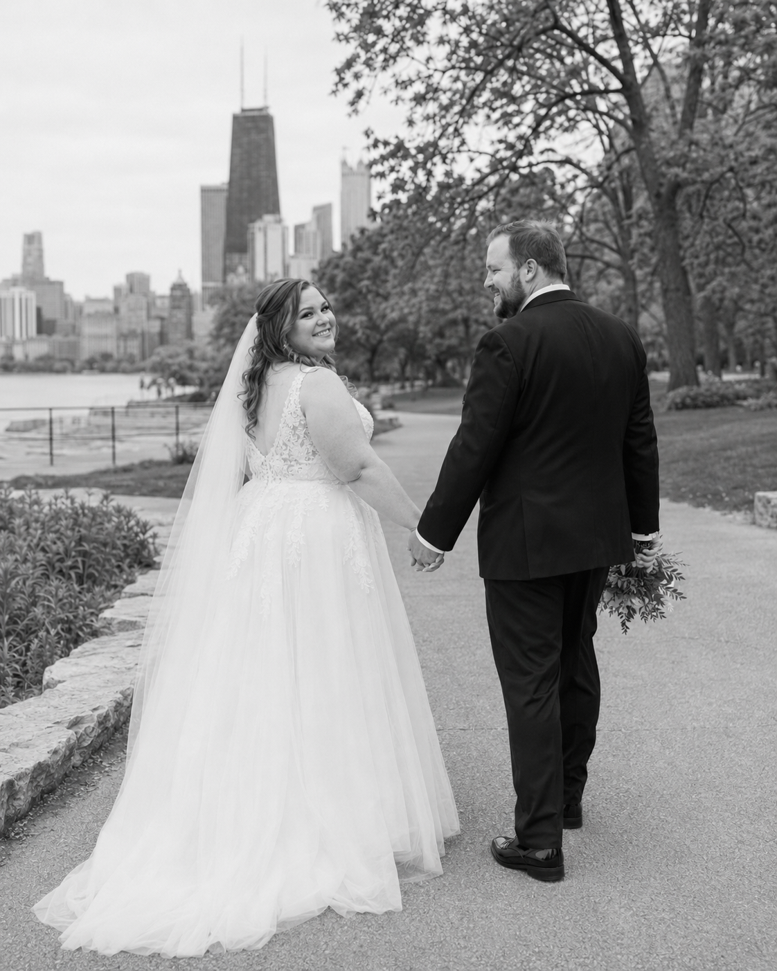 Black and white photo of bride and groom holding hands walking in park with Chicago skyline in background.