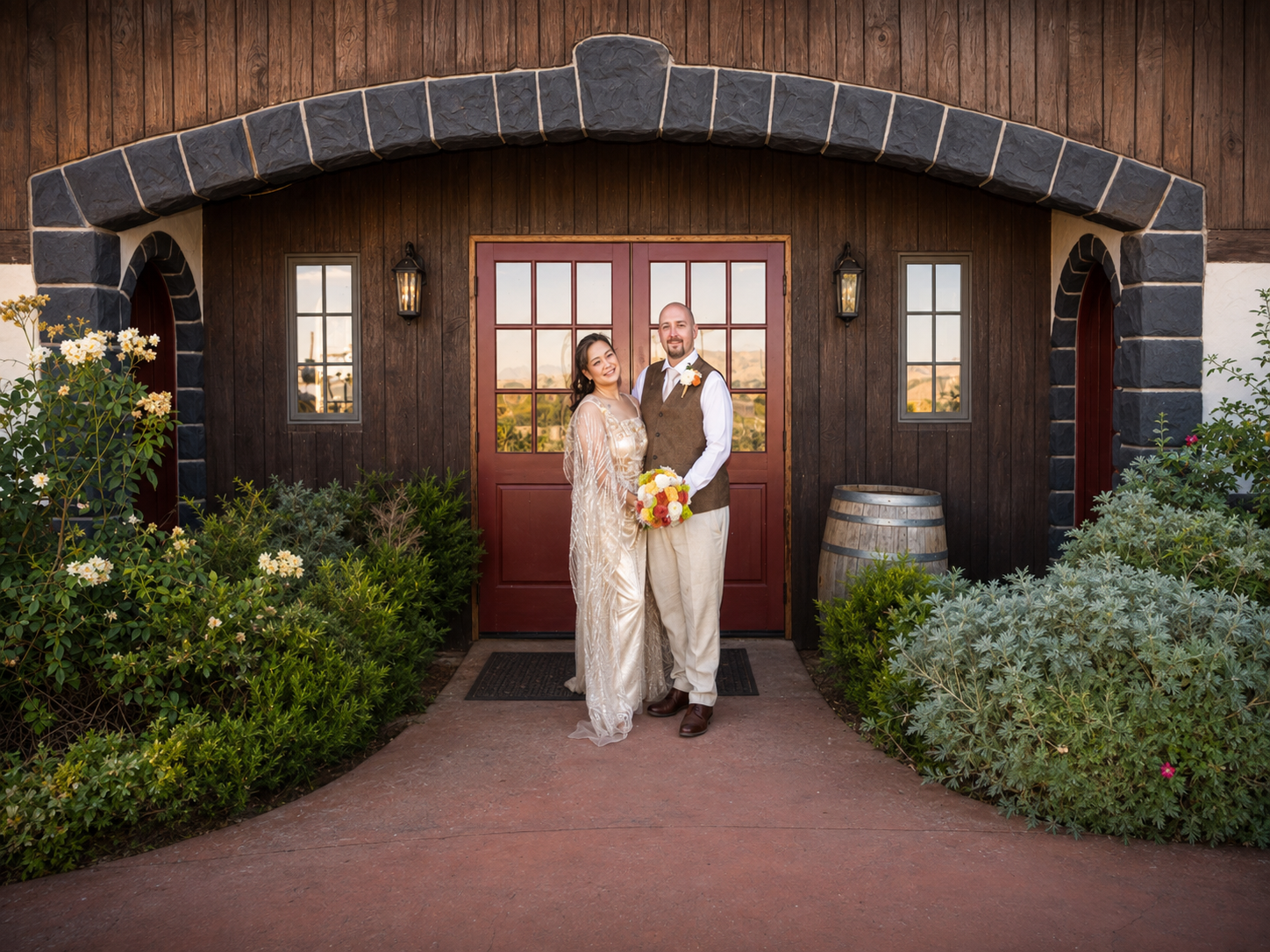 Bride and groom pose together in front of rustic wooden barn doors surrounded by lush greenery at winery wedding venue.