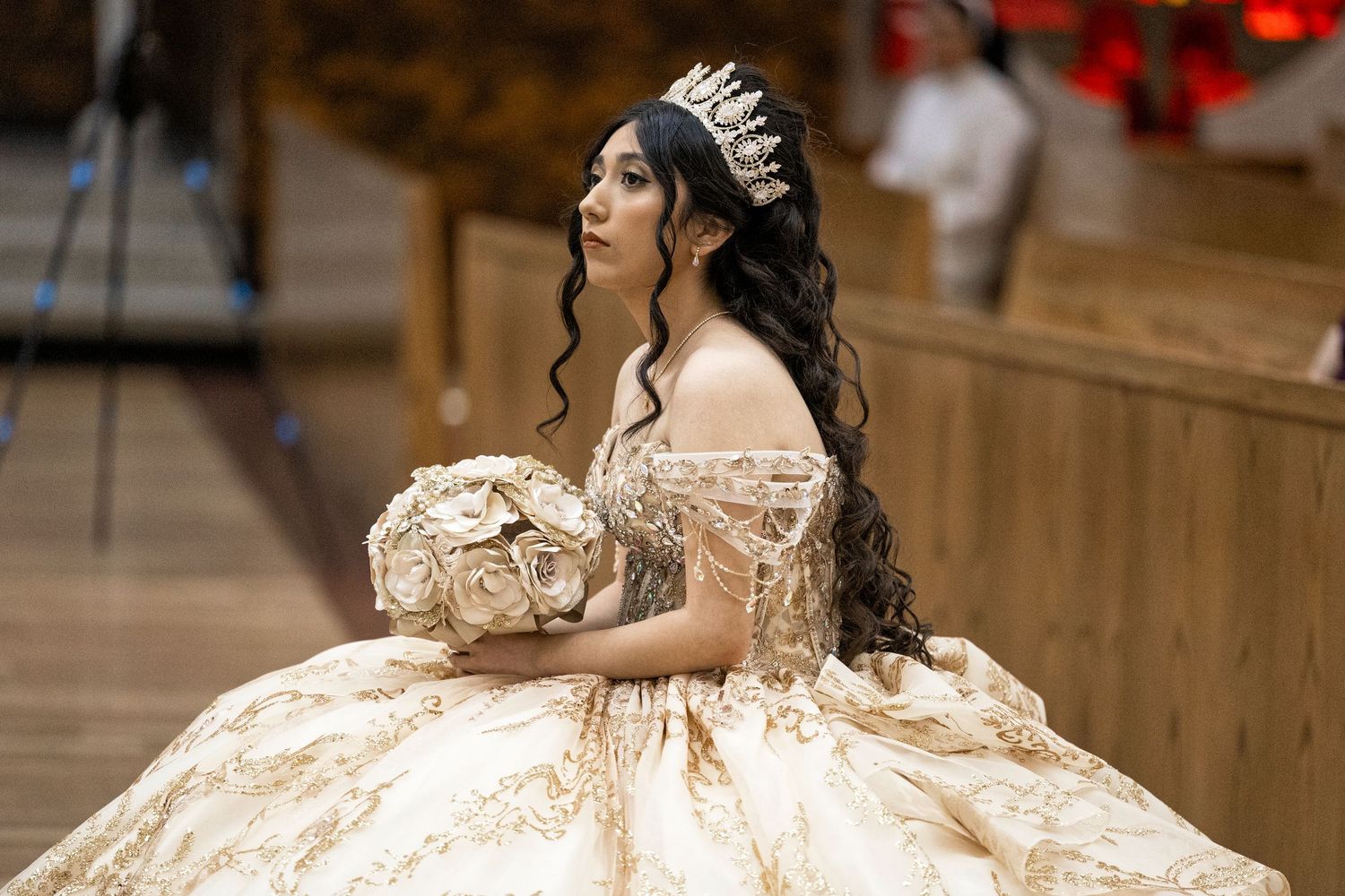 Young woman in ornate gold quinceañera ball gown with crown and floral bouquet poses elegantly in a grand venue.