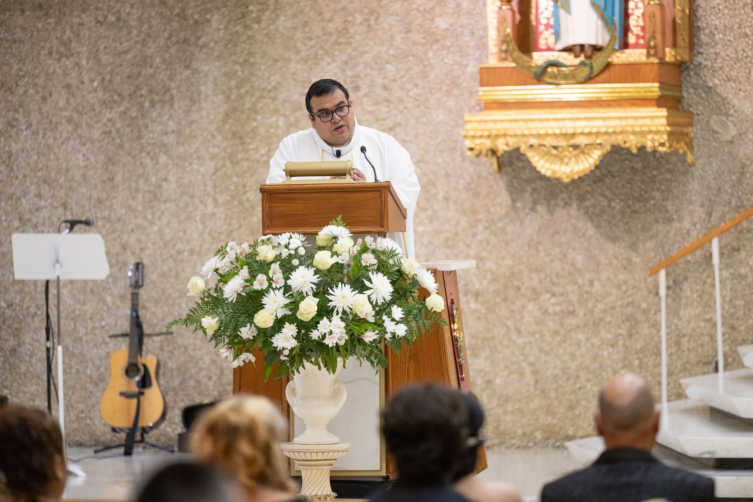 Priest delivering a sermon at a church podium adorned with white flowers during a religious ceremony.