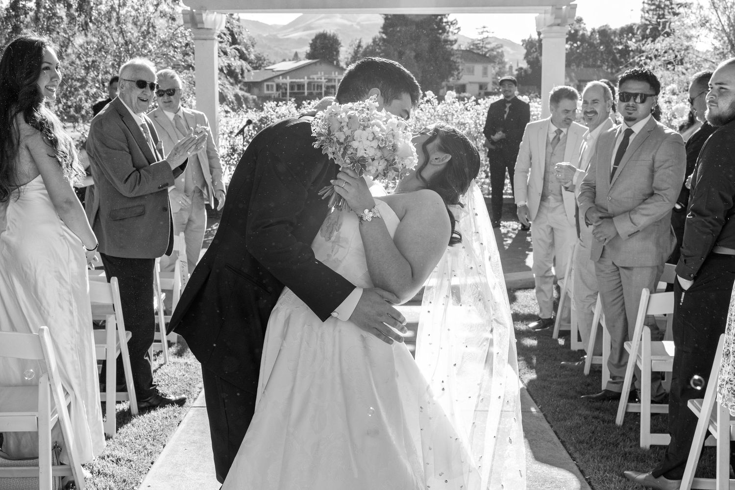 Black and white photo of a bride and groom sharing their first kiss at an outdoor wedding ceremony with guests watching.