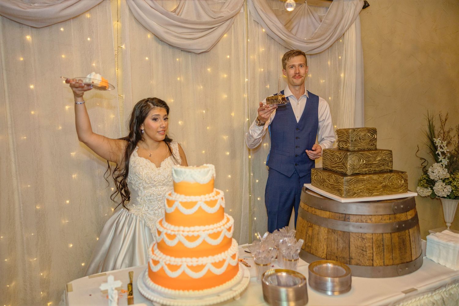 Bride and groom celebrating at wedding reception beside a three-tier orange cake on a decorated table.