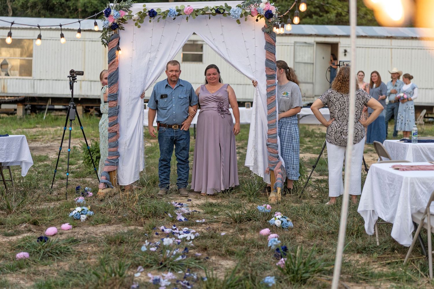 Couple stands under floral wedding arch outdoors at dusk, guests mingle nearby with decorated tables and string lights.
