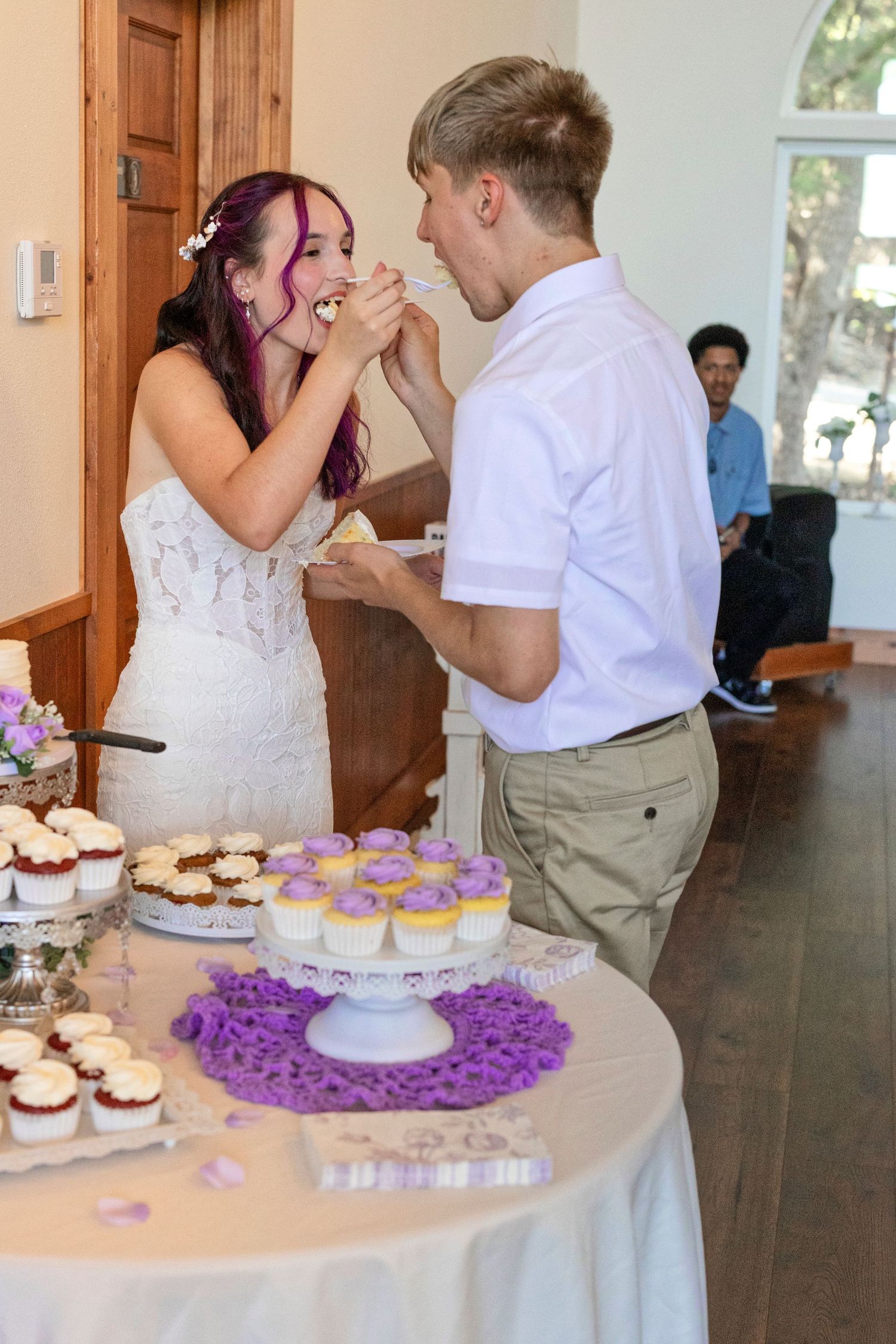 Bride and groom feeding each other wedding cake at reception table with purple cupcakes.