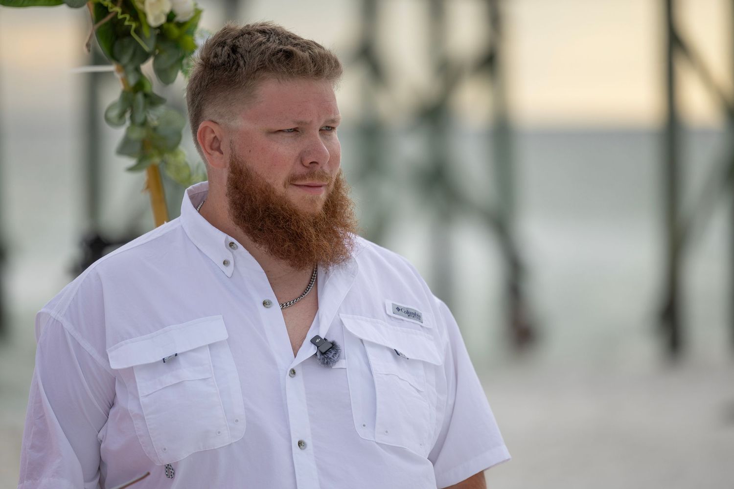 Man with red beard in white shirt standing on beach near pier at sunset outdoor wedding ceremony.