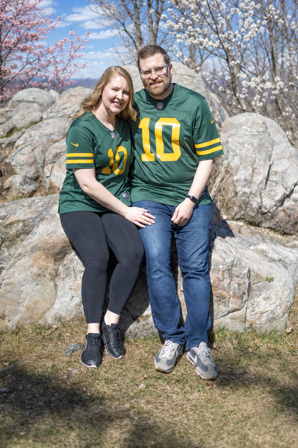 Couple wearing matching green number 10 football jerseys sitting on rocks with spring blossoms in background.