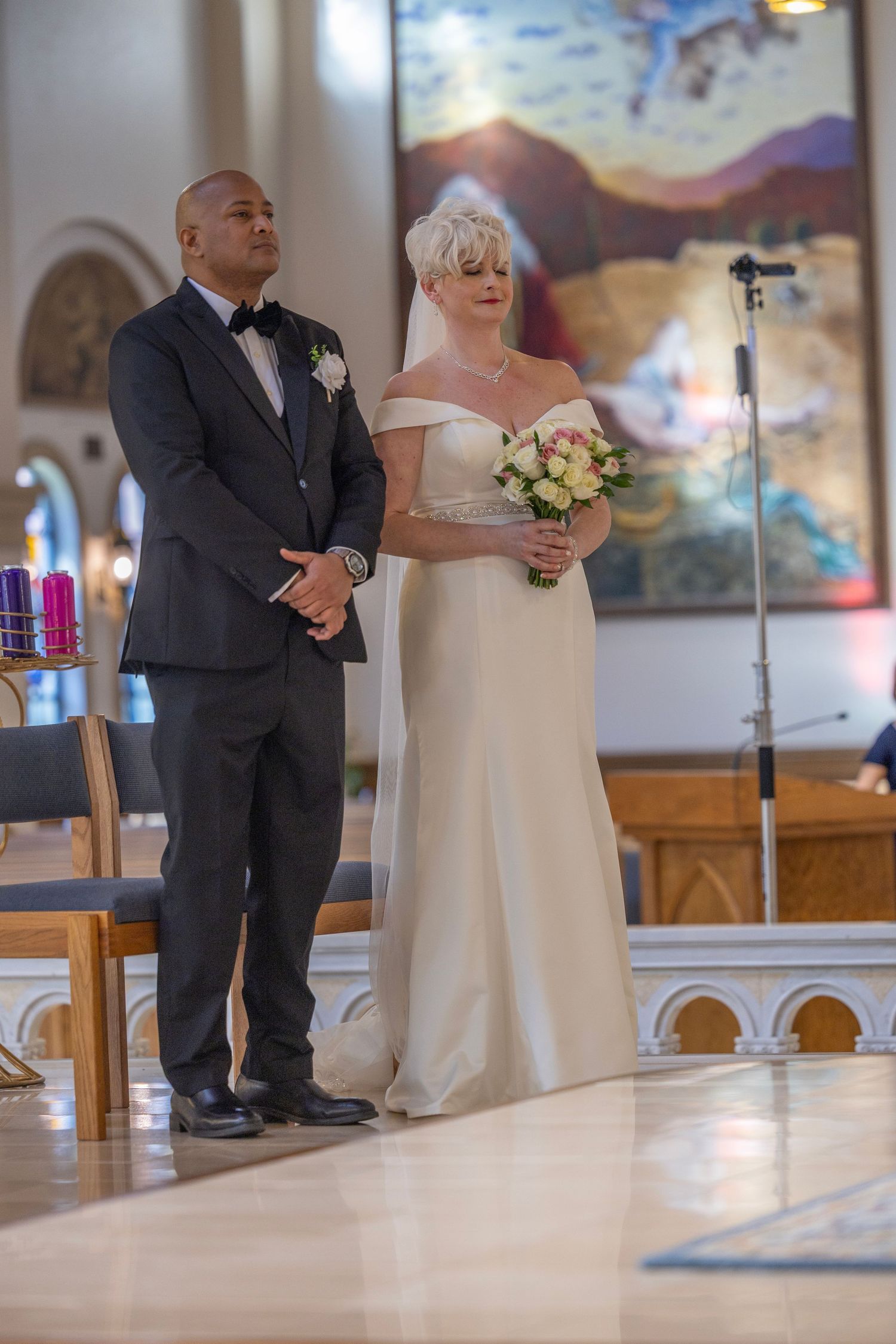 A bride in an off-shoulder white gown and groom in a black tuxedo stand at the altar of a church during their wedding ceremony.