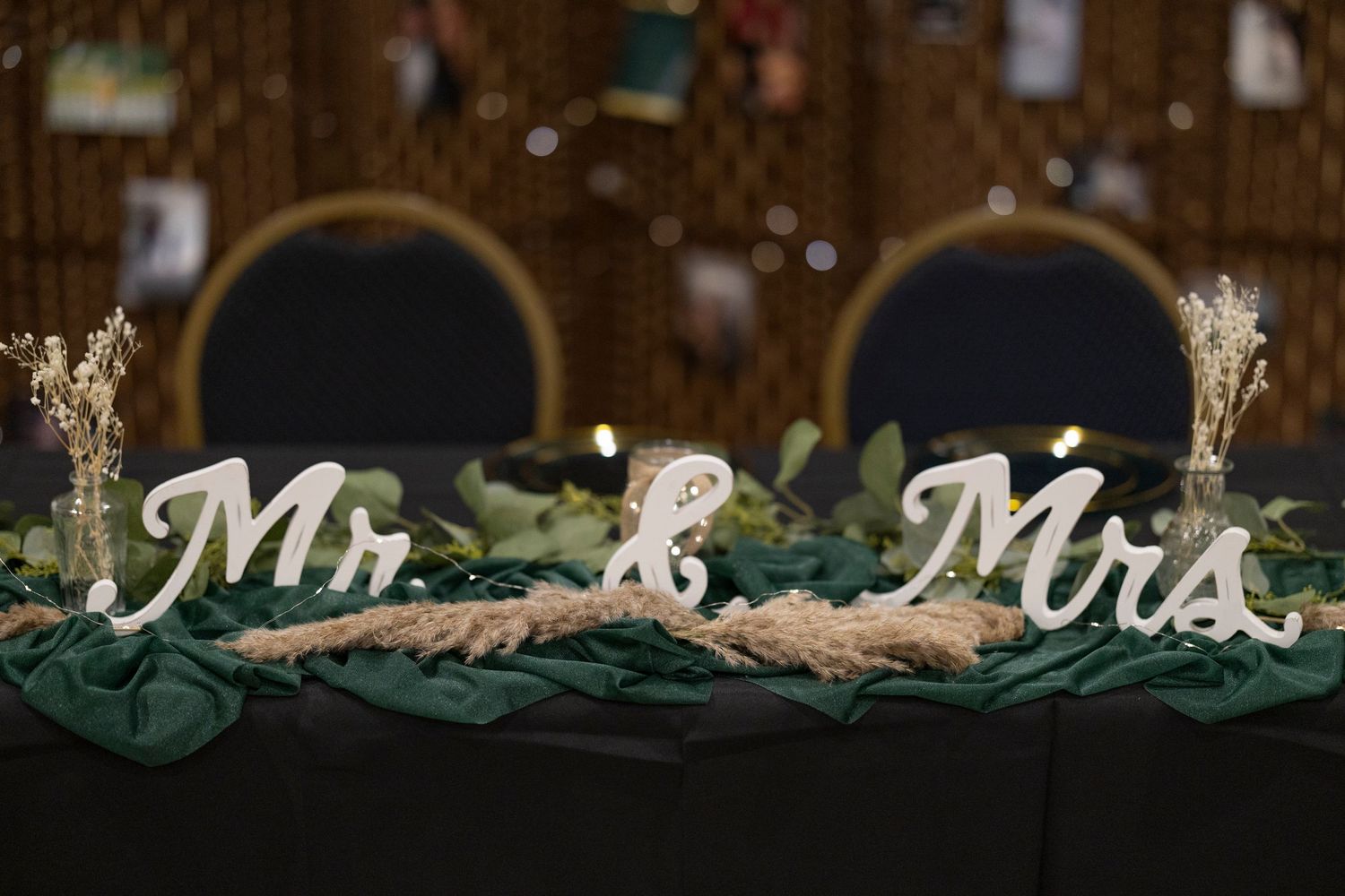 Rustic wedding decor featuring wooden Mr & Mrs signs with greenery and dried flowers on dark emerald fabric.