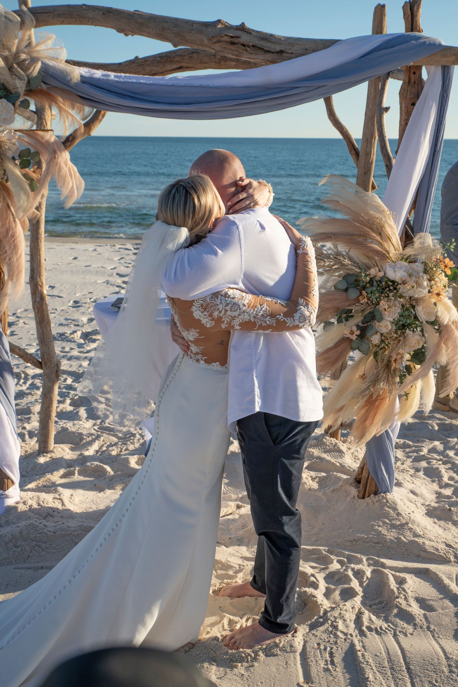 A romantic beach wedding ceremony takes place under a draped canopy with ocean waves in the background.