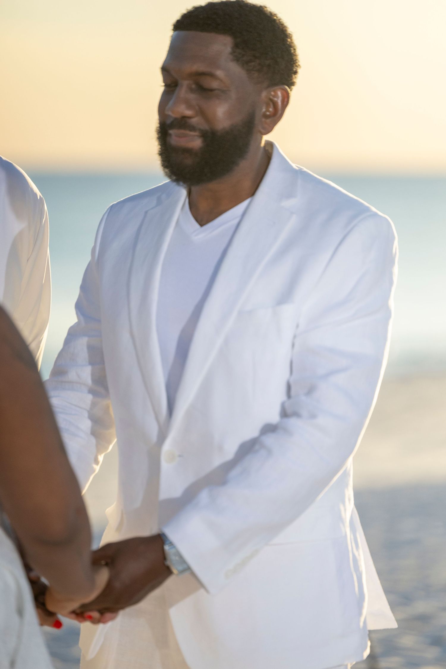 Two people in white attire sit together on a beach at sunset creating a romantic seaside moment.